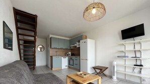 A compact living space with a kitchenette, a sofa, a coffee table, a TV mounted on the wall, and a ladder against the wall. - Renovated 2-bedroom apartment with pool and parking, sleeps 6 near the beach (Gruissan)