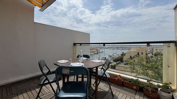 A balcony with a table set for two, chairs, potted plants, and a view of a marina and buildings. - Renovated 2-bedroom apartment with pool and parking, sleeps 6 near the beach (Gruissan)