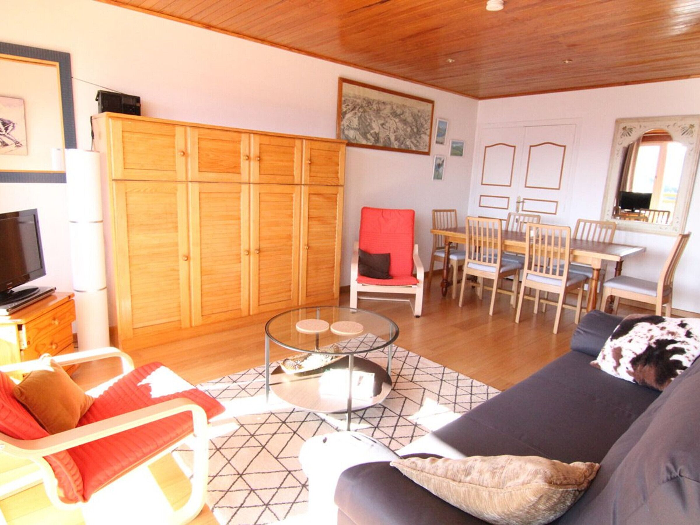 A living room with a sofa, a glass coffee table, a red chair, wooden cabinets, a TV, and a dining area with chairs and a table.