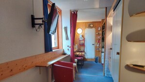 Ein schmaler Korridor mit einer hölzernen Bank, einem roten Stuhl, einem Bett mit roter Decke, einem kleinen Tisch und einer Tür zu einer Küchenzeile mit Regalen und einem Kühlschrank.