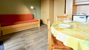 Un espacio habitable compacto con un sofá de madera, una mesa de comedor para dos personas y una kitchenette al fondo.