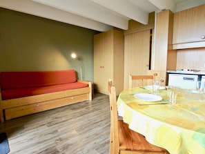 A compact living space with a wooden sofa, a dining table set for two, and a kitchenette in the background.
