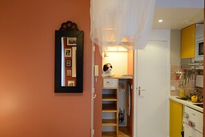 A narrow hallway with a mirror on the left, a small table with a stuffed dog, and a kitchen area with yellow cabinets and a stove.