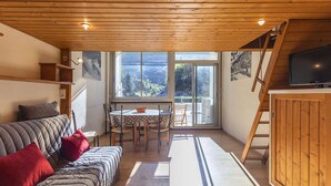 A cozy living room with a wooden ceiling, a sofa with red cushions, a dining table with chairs, and a TV on a wooden cabinet.