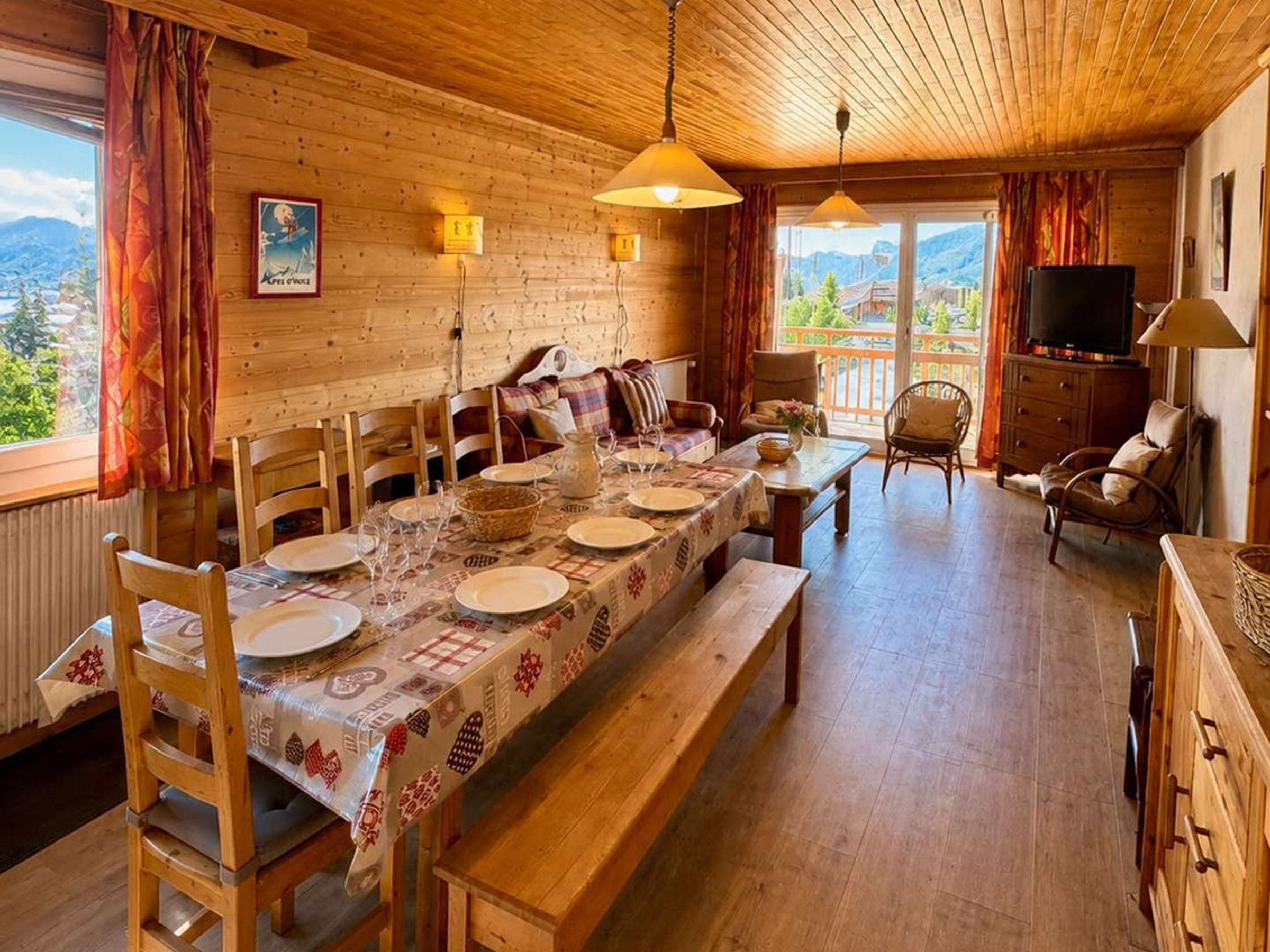 Un interior de madera con una mesa de comedor lista para cuatro personas, sillas de madera, un banco, un televisor y vistas al exterior.