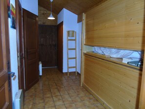 A narrow hallway with wooden walls and a tiled floor. A ladder is leaning against the wall, and a door is open at the end of the hallway.