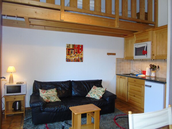 A living room with a black leather sofa, a small wooden table, a TV on a wooden stand, and a kitchen area with wooden cabinets and a microwave. - Spacious apartment for 6 with balcony in Châtel (Châtel)