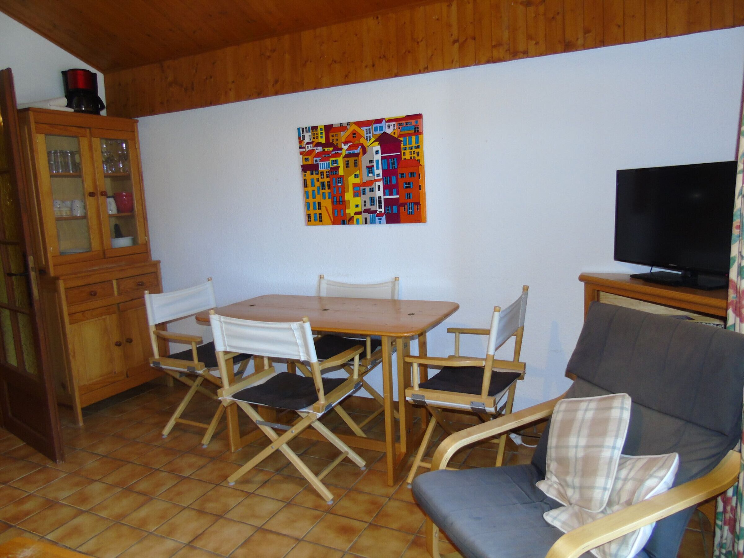 Uma área de refeições com uma mesa e cadeiras de madeira, um armário com portas de vidro, um quadro colorido na parede e uma televisão sobre um suporte.