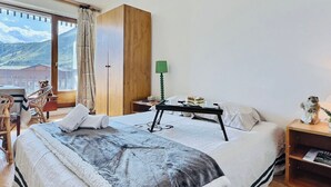 A bedroom with a bed, a wooden wardrobe, a nightstand with a lamp, a small table with a teddy bear, and a view of mountains through a window. - 2-room apartment, 6 ppl, Tignes, near slopes and shops (Tignes)