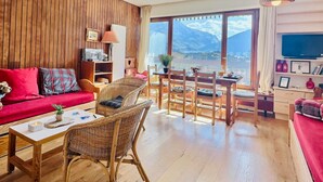 A cozy living room with a wooden wall, a red sofa, a dining area with a table and chairs, and a large window offering a mountain view. - 2-room apartment, 6 ppl, Tignes, near slopes and shops (Tignes)