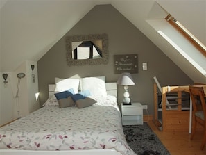 A bedroom with a sloped ceiling, a bed with patterned bedding, a bedside lamp, a small desk, and a mirror on the wall.