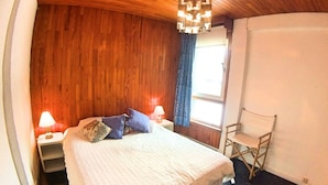 Un dormitorio con techo de madera, una cama con colcha blanca, dos lámparas de noche, una ventana con cortinas y una silla.