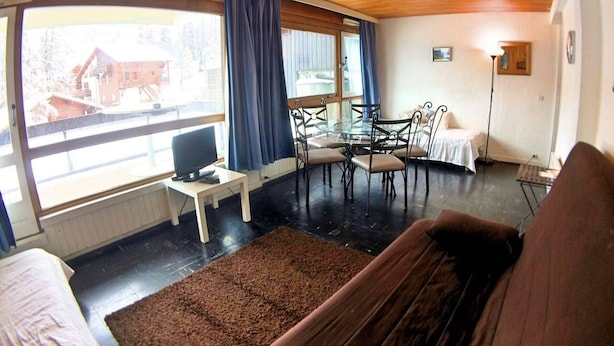Una habitación acogedora con techo de madera, una mesa redonda de vidrio con sillas, un televisor sobre una mesita y un ventanal con cortinas.