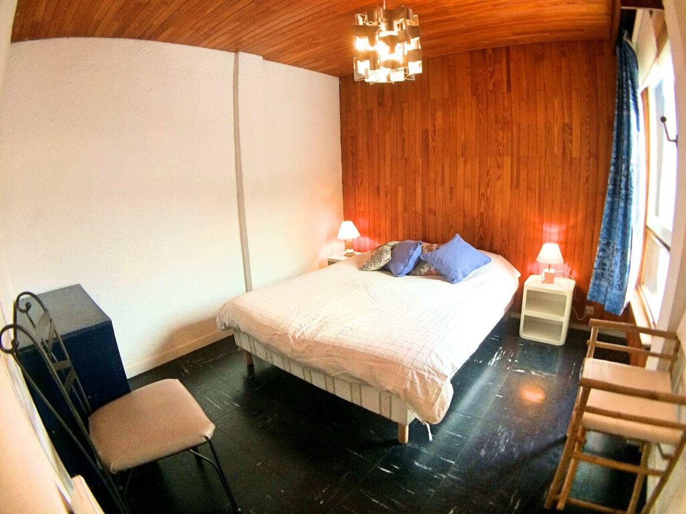 A bedroom with a wooden ceiling, a bed with pillows, a nightstand with a lamp, a chair, and a desk.
