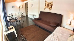 Una habitación moderna con un sofá marrón, una cama con cubierta blanca, una zona de comedor con sillas y una mesa, y un reloj fijado a la pared.