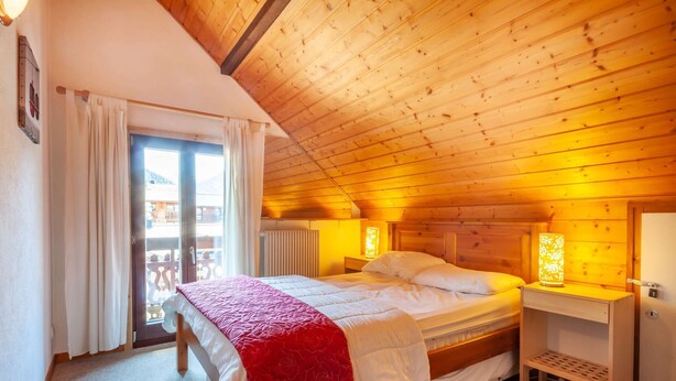 A cozy bedroom with a wooden ceiling, a bed with a red and white bedspread, a nightstand with a lamp, and a view of the outside balcony.