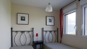 Une chambre avec deux lits simples, une petite table surmontée d’une lampe rouge et deux tableaux encadrés accrochés au mur.
