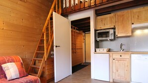Une cuisine compacte avec des meubles en bois, un four à micro-ondes et un petit réfrigérateur. Une échelle en bois mène à une mezzanine aménagée en espace nuit avec un lit.