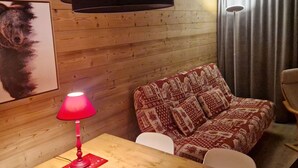 A cozy room with wooden walls, a patterned sofa, a red table lamp, and a framed picture on the wall. - Cozy studio for 4 in L'Alpe d'Huez, near slopes, pets welcome (Huez)