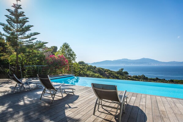 A wooden deck with lounge chairs overlooking a clear blue pool and a scenic view of the sea and mountains.