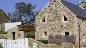 A stone building with a sloped roof, a wooden fence, and a small garden area. - Cozy Cottage with Fireplace, Garden, and Game Room near the Sea (La Hague)