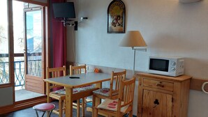 Una habitación con una mesa de madera, cuatro sillas, un televisor montado en la pared, un microondas sobre un mueble de madera y una ventana con vistas a árboles.