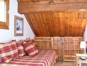 Une chambre mansardée chaleureuse avec un plafond en bois, un lit recouvert d’une couette à carreaux et une petite table sur laquelle se trouvent une lampe et une télécommande.
