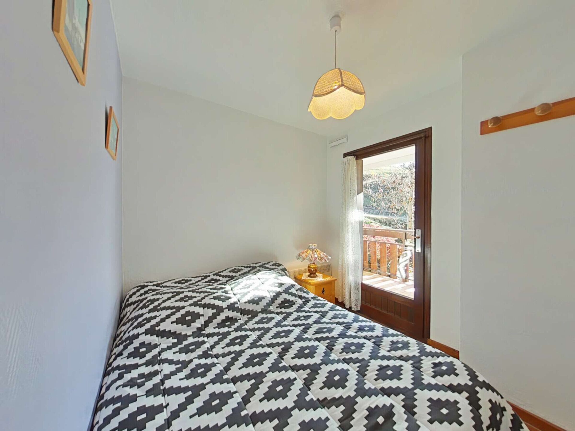 Ein Schlafzimmer mit einem gemusterten Bett, einer Nachttischlampe und Blick auf einen Balkon mit Geländer.