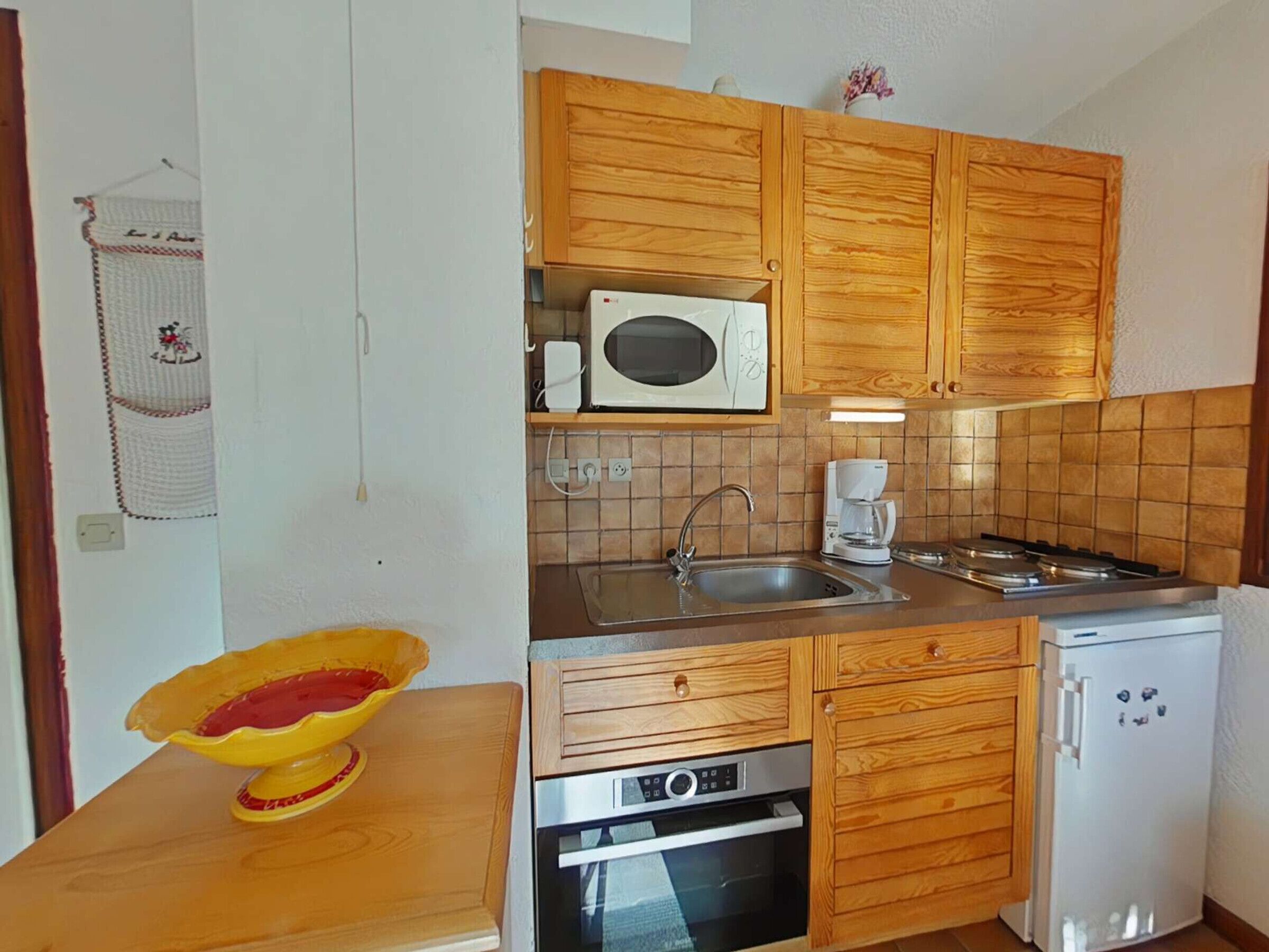 Una cocina compacta con armarios de madera, microondas, cafetera, pileta, estufa y un refrigerador pequeño.
