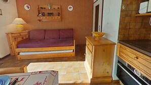 Une pièce de taille modeste, comprenant un lit en bois, une table de chevet avec une lampe, une petite étagère avec des objets décoratifs et un coin cuisine équipé d’une cuisinière.