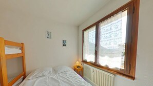 Un lit simple avec une couette blanche, un lit superposé en bois sur la gauche, une petite table de chevet avec une lampe et une fenêtre avec des rideaux à motifs.