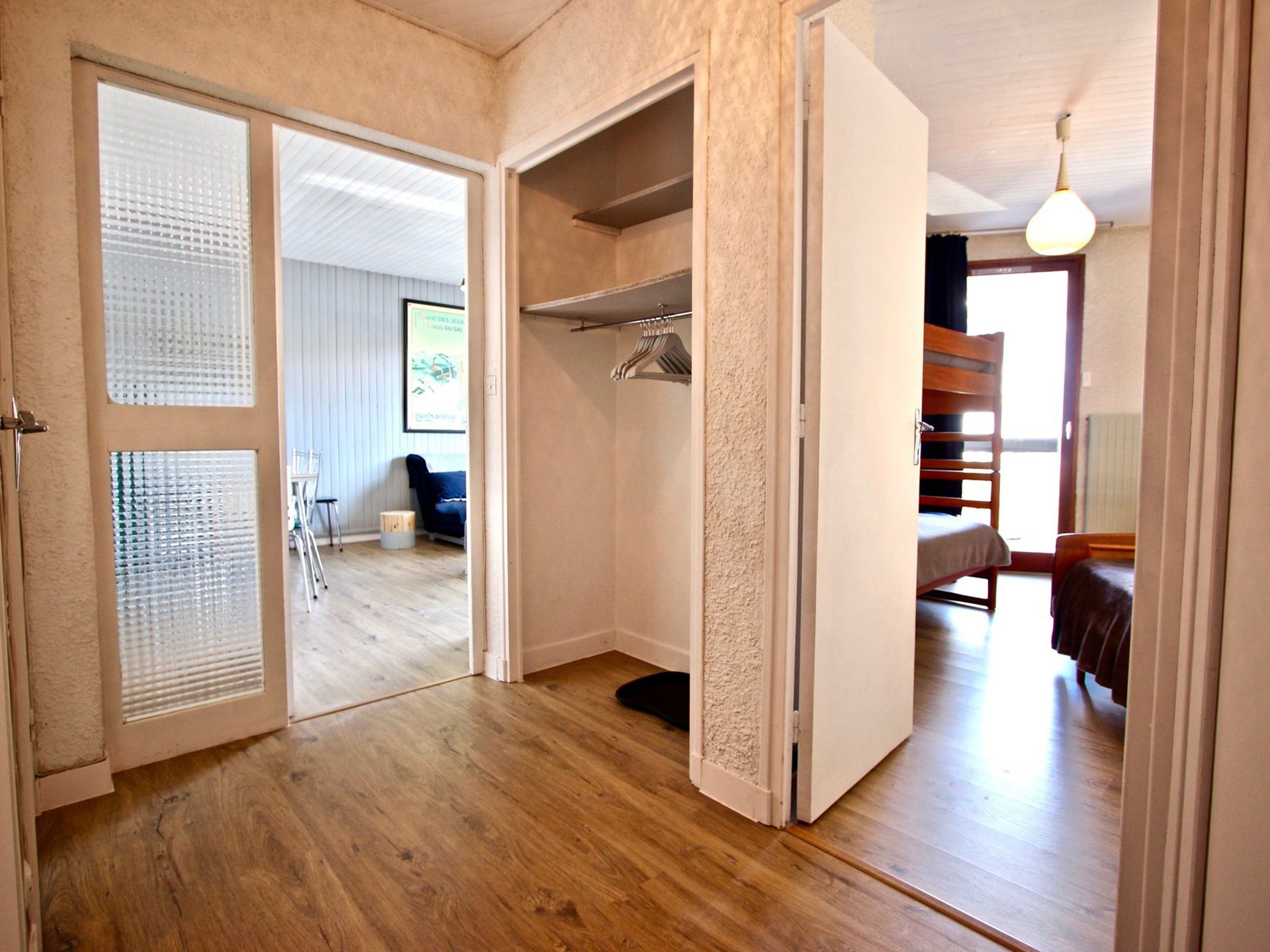 A room with a wooden floor, a bed, a wardrobe, and a door leading to another room.