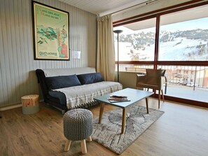 A cozy room with a sofa, a small table, and a chair. There's a poster on the wall and a window with a view of a snowy landscape.