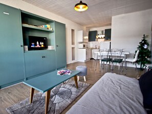 A modern living room with a teal cabinet, a dining area with a white table and chairs, and a kitchen in the background.