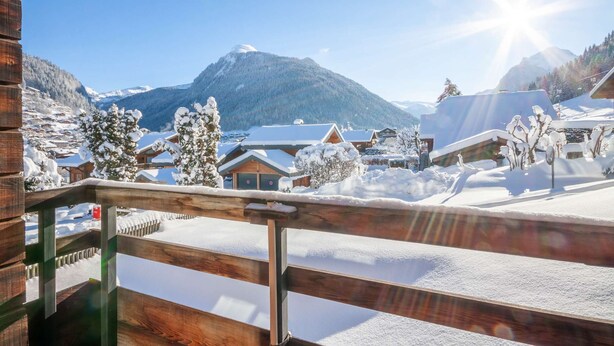 Ein Holzbalkon mit verschneiter Landschaft, einschließlich Häusern und Bergen im Hintergrund.