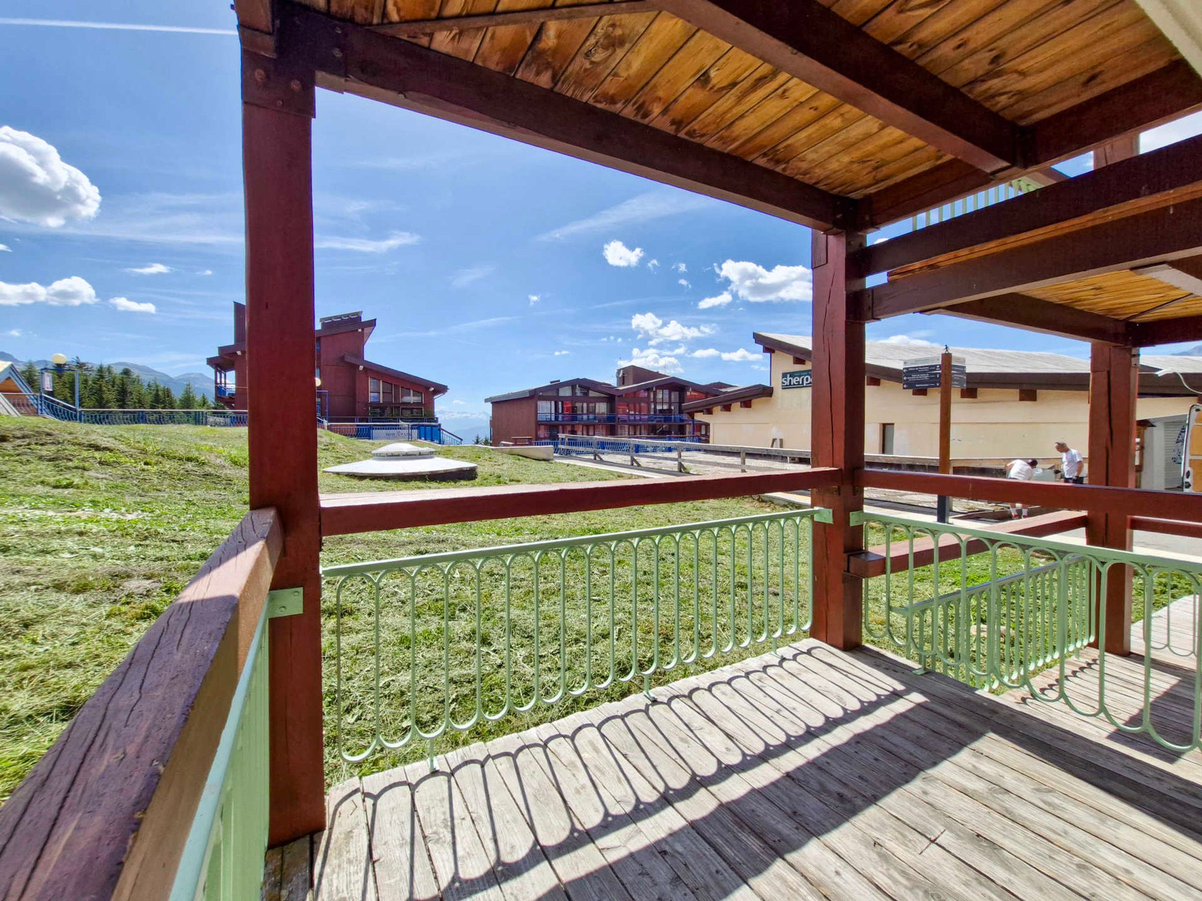 A wooden deck with a metal railing overlooking a grassy area with buildings in the background.