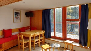 Quarto aconchegante com teto de madeira, um sofá com almofadas vermelhas, uma mesa de madeira e cadeiras. Há uma TV na parede e uma janela com cortinas.