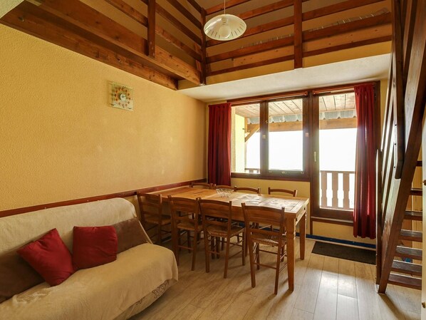 A dining area with a wooden table and chairs, a sofa with red cushions, and a window with curtains.