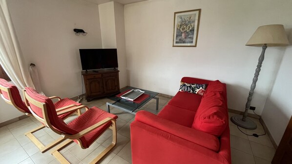 A living room with a red sofa, two red chairs, a glass coffee table, a TV on a wooden cabinet, a floor lamp, and a framed picture on the wall.