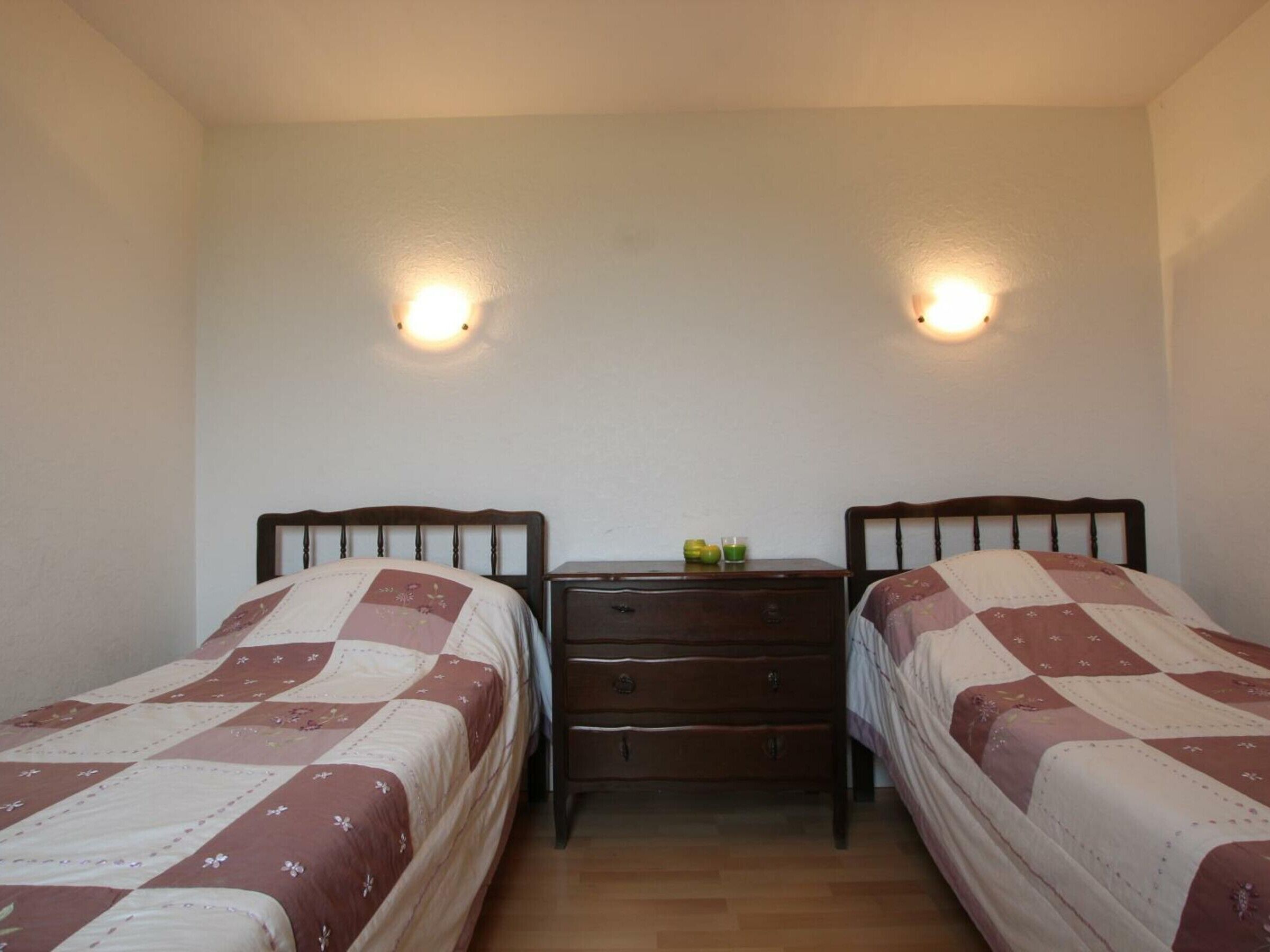 Una habitación pequeña y limpia con dos camas individuales, una mesita de noche entre ellas y dos lámparas de pared.