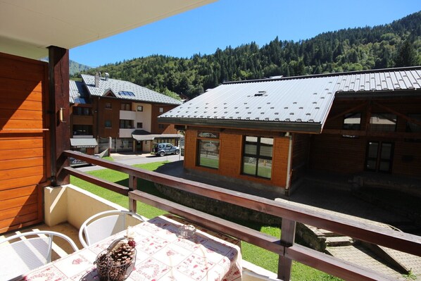A wooden balcony with a table and chairs, overlooking a building with a metal roof and a view of a forested mountain.