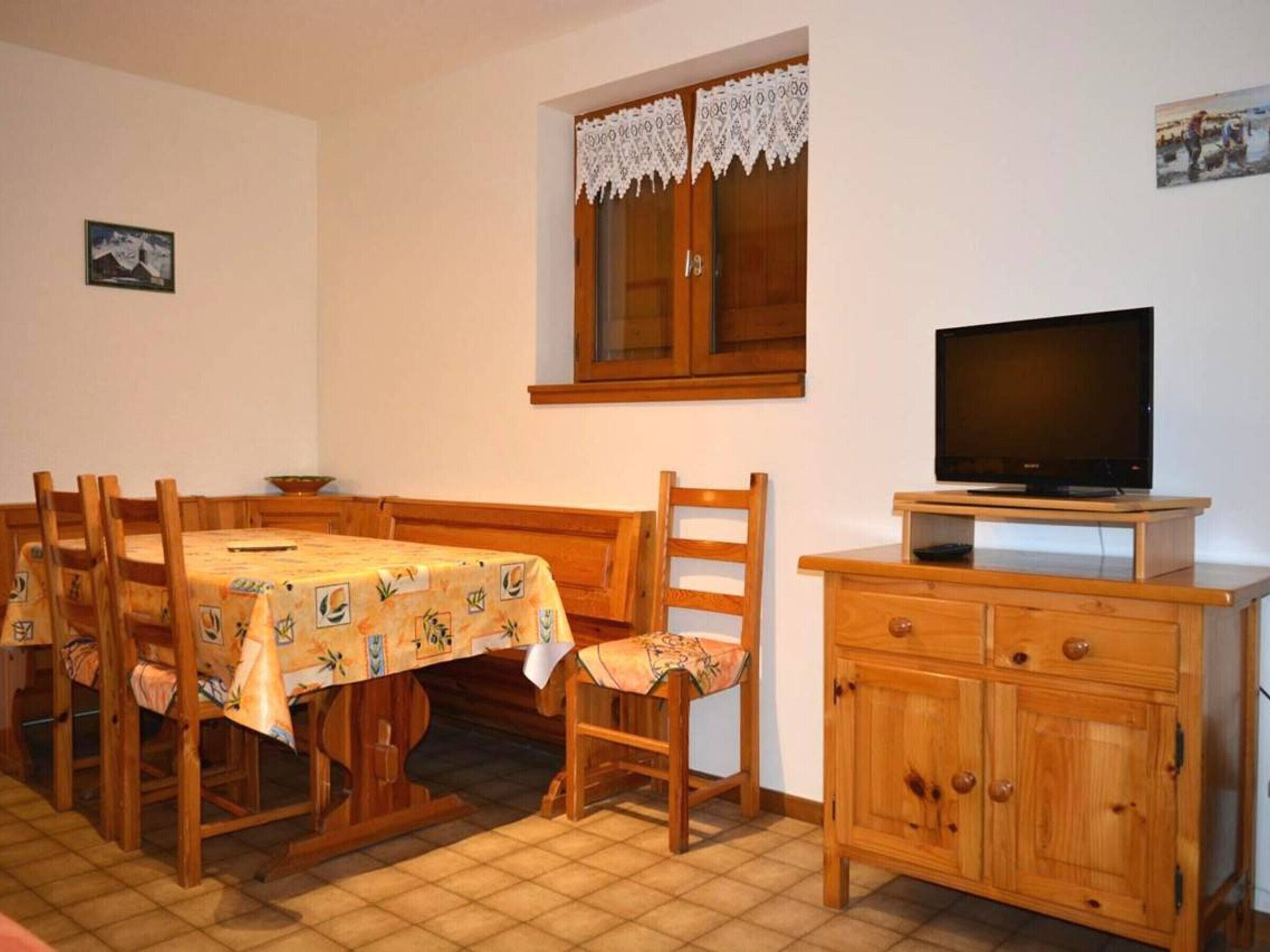 Ein Essbereich mit Holztisch und -stühlen, einem Fernseher auf einem Holzschrank und einem Fenster mit Spitzenvorhängen.