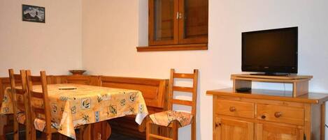 Ein Essbereich mit Holztisch und -stühlen, einem Fernseher auf einem Holzschrank und einem Fenster mit Spitzenvorhängen.