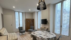 A modern living room with a sofa, two armchairs, a dining table with a newspaper-covered tablecloth, a wooden cabinet, a television on a stand, and large windows with frosted glass. - T2 Apartment for 5 in Cauterets (Cauterets)