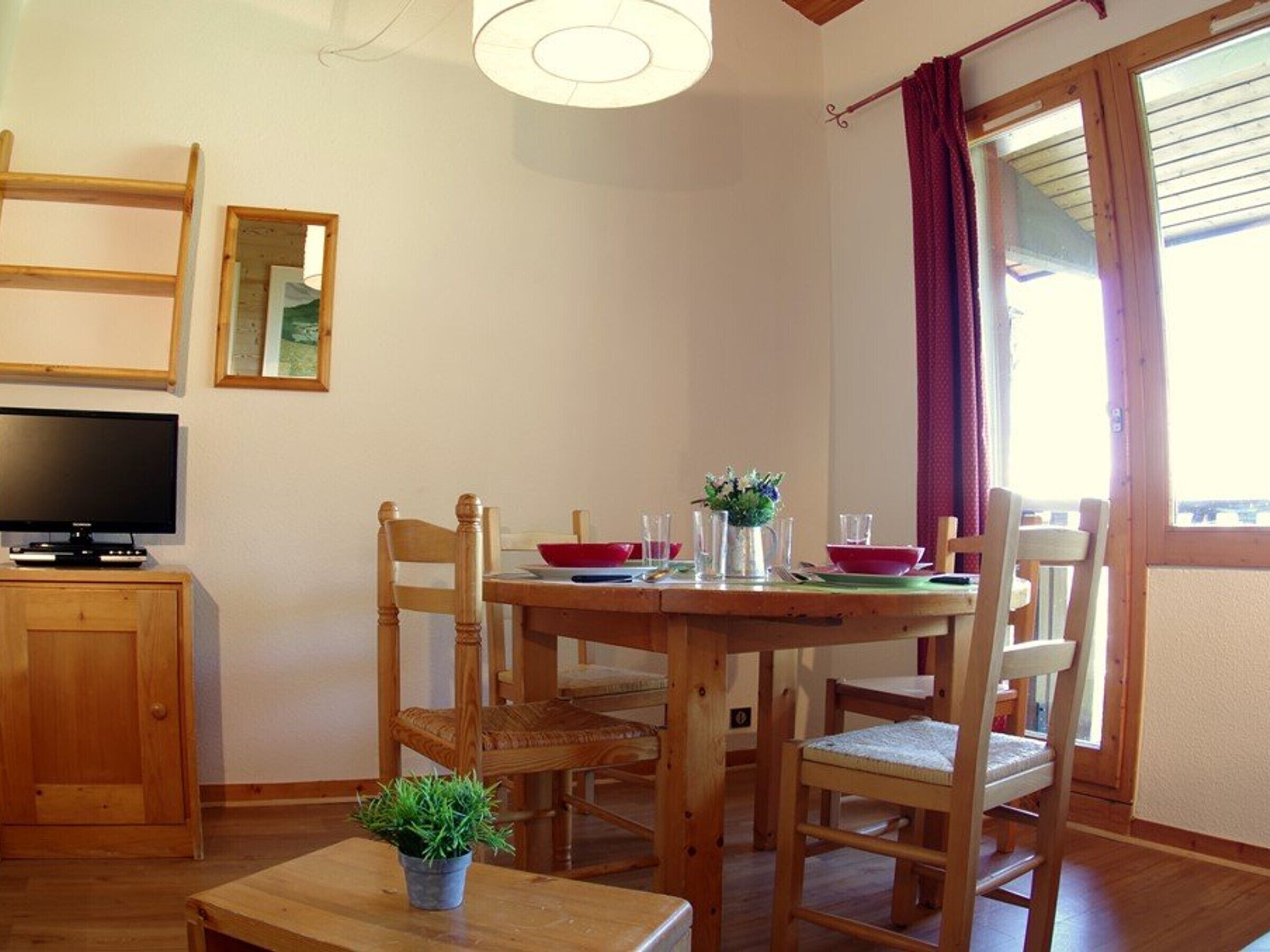 A dining area with a wooden table, chairs, a television, and a window with curtains.