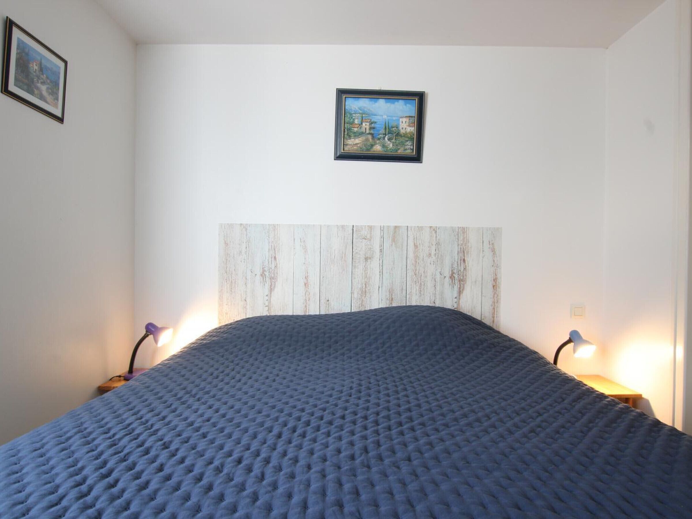 A bedroom with a wooden headboard, a blue textured bedspread, two bedside lamps, and a framed picture on the wall.