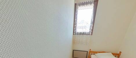 A small, narrow room with a wooden floor, a single bed with white bedding, a radiator, and a window with lace curtains.