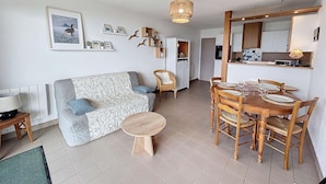 A living room with a sofa, a dining table set for four, and a kitchen in the background. - Beachfront Apartment with Terrace, Stunning Sea View, Parking & WiFi in Jullouville (Jullouville)