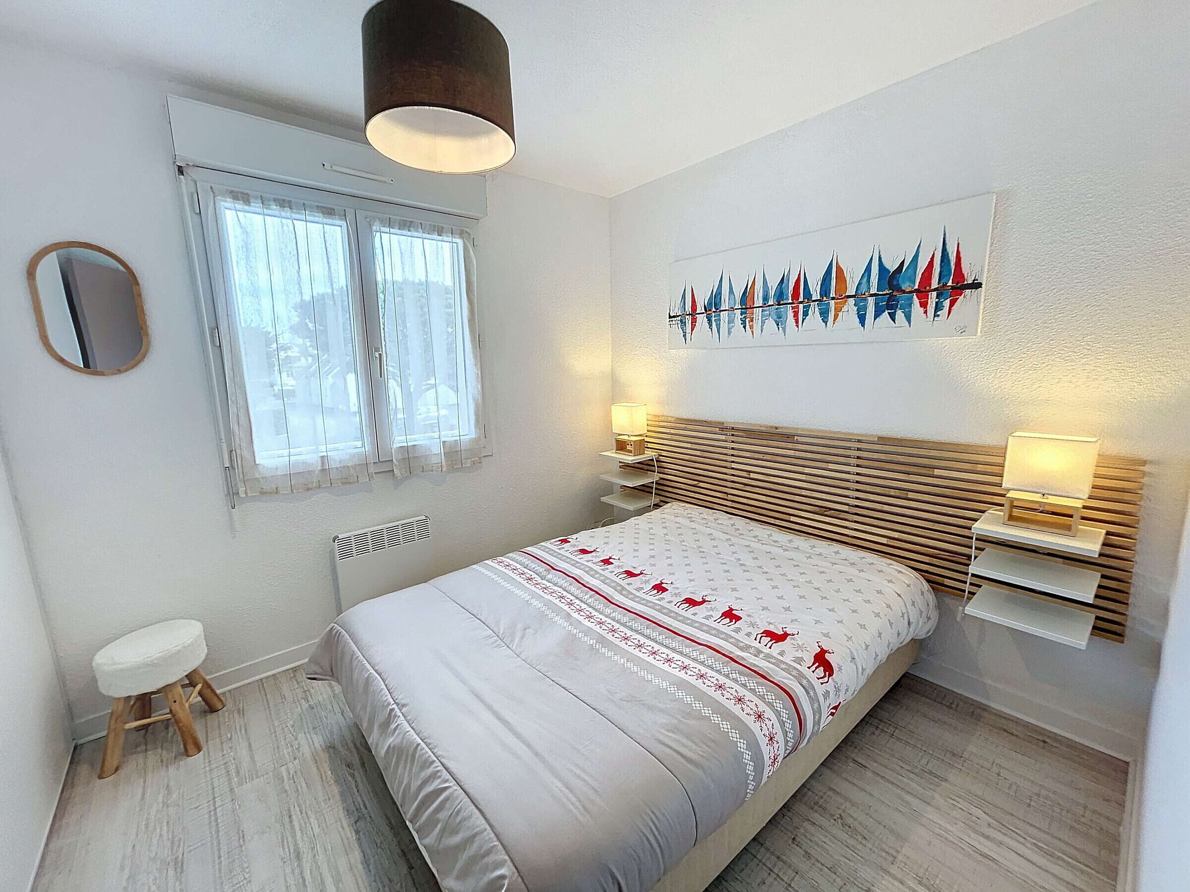 Un dormitorio con cama, lámparas de noche, ventana con cortinas, radiador, un taburete pequeño y estante fijado a la pared.