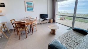 A modern living room with a dining area, a television, and a large window overlooking a beach. - Beachfront Apartment with Terrace, Stunning Sea View, Parking & WiFi in Jullouville (Jullouville)